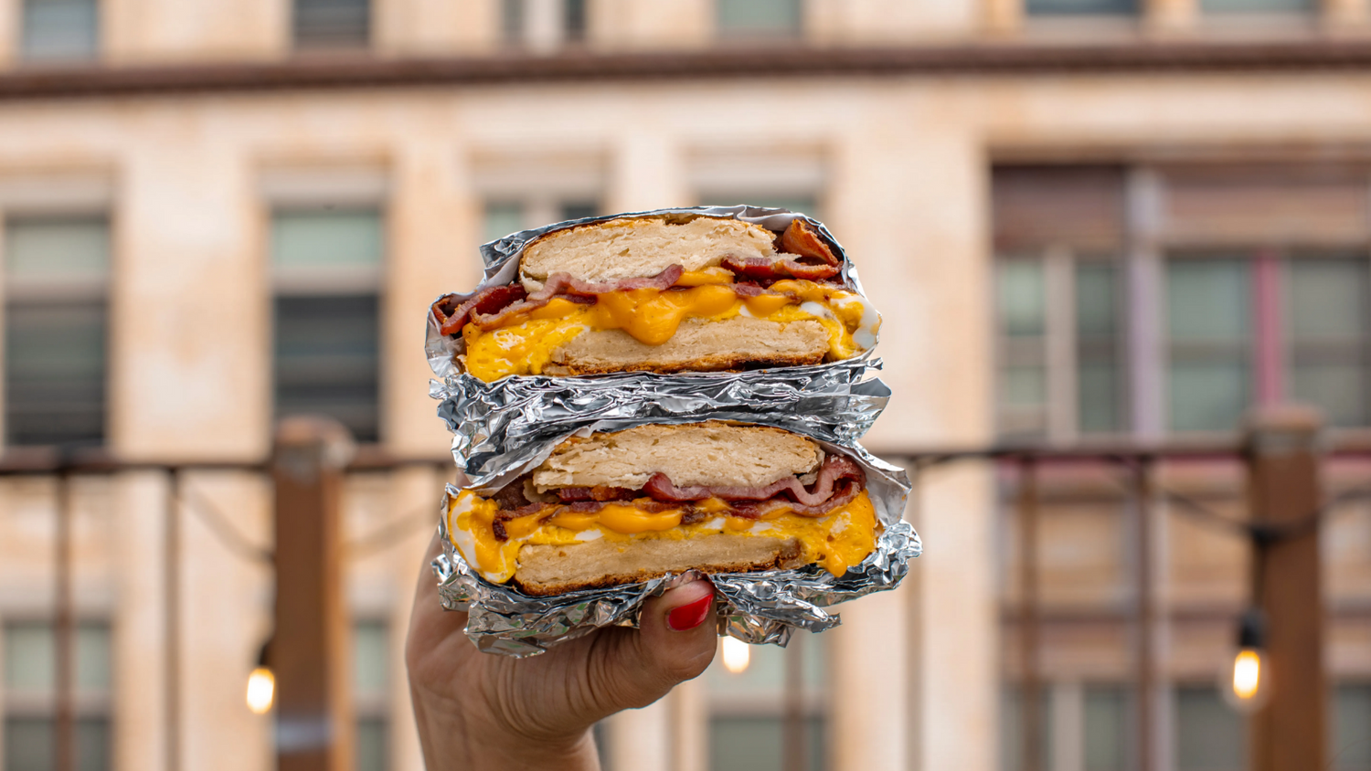 bacon, egg, and cheese breakfast sandwich in a persons hand with the city in the background